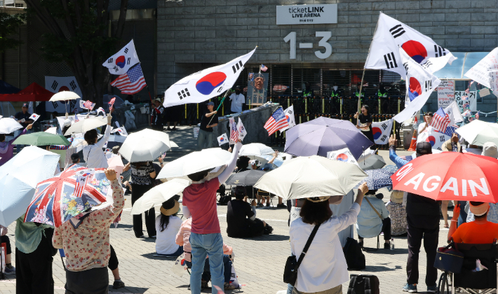 낮 최고 기온이 29도로 예보된 23일 6·3 지방선거 잠실 개표소로 사용된 서울 송파구 올림픽공원 핸드볼경기장 앞에서 투표용지 부족 사태 규탄 봉쇄 시위 참가자들이 우산을 쓰고 구호를 외치고 있다. 연합뉴스